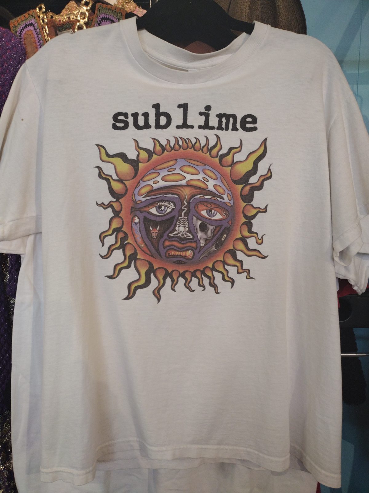 White t-shirt with black text "sublime" above a colorful sun graphic with a face.